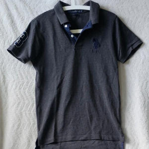Mörkgrå pikétröja från U.S. Polo Assn. - Klassisk mörkgrå pikétröja från U.S. Polo Assn. med broderad logga på bröstet och nummerdetalj på ärmen. Tröjan har korta ärmar, krage och knappar framtill. Perfekt till jeans eller shorts för en avslappnad look.