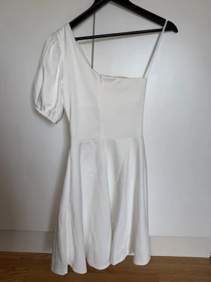 Vit oneshoulder klänning - Vit klänning med oneshoulder-design och en puffärm. Klänningen har en figurnära överdel och utställd kjol. Fin på studenten!
