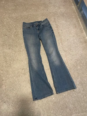 Blå bootcut jeans från vero Moda  - Low waist Snygga blå bootcut jeans med klassisk femficksdesign och utsvängda ben. Jeansen har en ljus tvätt och subtila slitningar framtill för en avslappnad look. Perfekta för dig som gillar  och vill ha ett par jeans som sticker ut.liten  vid foten nere på jeansen 