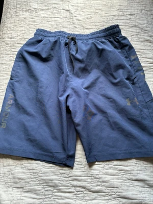 Blå träningsshorts från Under Armour - Säljer ett par blå träningsshorts från Under Armour med snörning i midjan och logga på benet. Shortsen har normal passform och är perfekta för träning. Tillverkade i ett mjukt och lätt material som känns skönt mot huden.