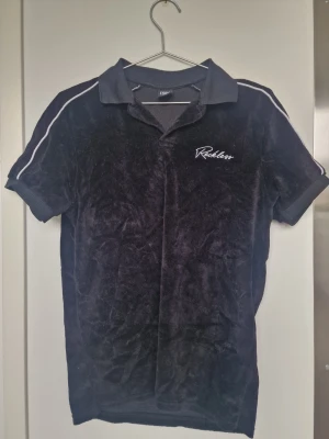 Svart polotopp från Reckless - Svart polotopp från Reckless med korta ärmar och vit piping längs axlarna. Mjuk och lite glansig yta, troligen i polyester. Broderad logga på bröstet och klassisk krage. Perfekt för en sportig och avslappnad look.
