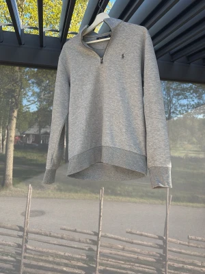 Grå halvzip tröja från Polo Ralph Lauren - Snygg grå sweatshirt med halv dragkedja från Polo Ralph Lauren. Tröjan har en liten broderad logga på bröstet, ribbade muddar och en avslappnad passform. Perfekt till jeans eller mjukisbyxor för en clean och enkel stil.