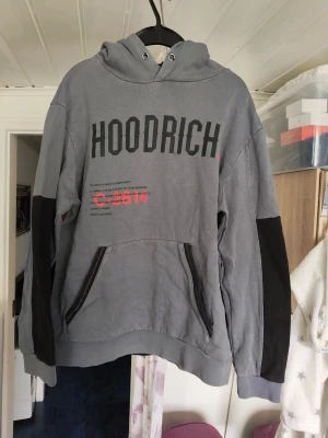 Grå hoodie från Hoodrich med tryck - Snygg grå hoodie från Hoodrich med stor svart logga på bröstet och detaljer i rött. Hoodien har huva, dragkedjor på fickorna och svarta paneler på ärmarna. Perfekt för en streetwear-look och riktigt bekväm med mjukt material. Saknar snöret vid huvan 