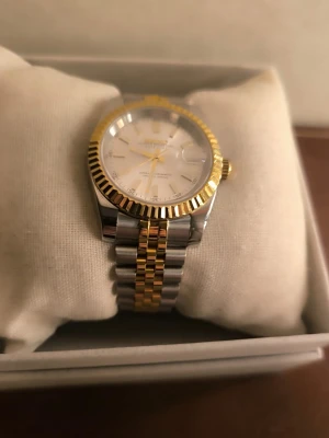 Seiko Mod Datejust tvåtonad klocka - Säljer en Seiko mod/custom i Datejust-inspirerad stil med tvåtonsutförande, räfflad guldfärgad bezel, jubilee-länk och champagne/silverfärgad tavla. Klockan har Seiko 4R35B automatiskt urverk, 23 jewels, med handuppdrag och hacking. Datumvisning med förstoringsglas, guldfärgade detaljer och glasbaksida. Mycket trevlig dress/sport-klocka med klassisk look. Passar bra om man gillar Datejust-stilen men vill ha något mer prisvärt och bärbart. Fint begagnat skick. Mindre bruksspår på spännet, se bild