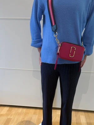 Rosa axelväska från Marc Jacobs - Cool axelväska från Marc Jacobs i stark rosa färg med bred axelrem i svart och rosa. Väskan har guldfärgade detaljer och ett stort metallspänne på framsidan. Materialet är läder och väskan har dubbla dragkedjor upptill.