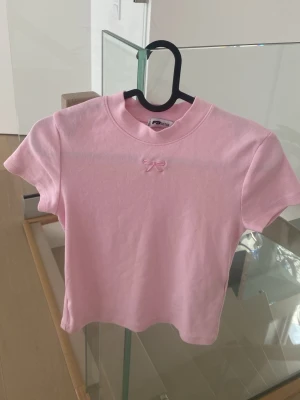 Rosa croppad t-shirt med rosett - Söt ljusrosa croppad t-shirt från Fruit of the Loom med en liten rosett framtill vid halsen. T-shirten har korta ärmar och är gjord i mjuk bomull, perfekt för en trendig och avslappnad look.
