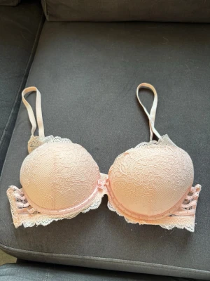Ljusrosa push-up bh med spets o pärlor  - Säljer en ljusrosa push-up bh med vadderade kupor, spetsdetaljer och dekorativa band med små nitar på sidorna. Bh:n har justerbara axelband och en söt rosett framtill. Perfekt för dig som gillar romantiska detaljer och mjuka pastellfärger. Storlek 75b lapp avklippt. Använd en gång