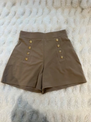 Beige högmidjade shorts med guldknappar - Stilrena beige shorts med hög midja och dekorativa guldknappar framtill i dubbla rader. Klassisk och trendig design som passar perfekt till sommaren. Mjukt material och snygg passform.