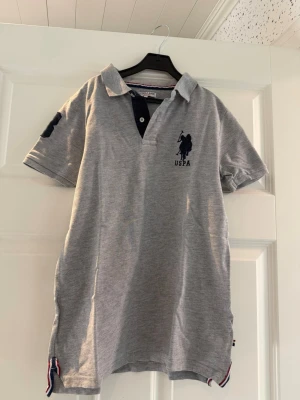 Grå pikétröja från U.S Polo Assn. - Grå pikétröja från USPA med klassisk krage, korta ärmar och mörkblå logga på bröstet. Tröjan har knappslå framtill och dekorativa banddetaljer i nederkant. Tillverkad i mjuk bomull för en bekväm känsla.