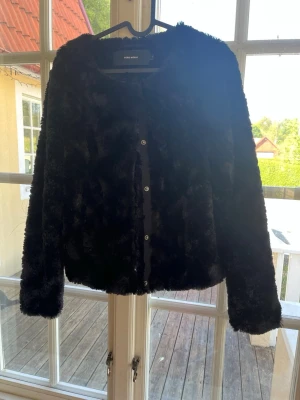 Svart teddyjacka från Vero Moda - Mysig svart teddyjacka från Vero Moda med rund halsringning och knäppning framtill. Jackan har en fluffig, mjuk yta i fuskpäls och raka ärmar. Perfekt för dig som vill ha en trendig och varm look under kyliga dagar.
