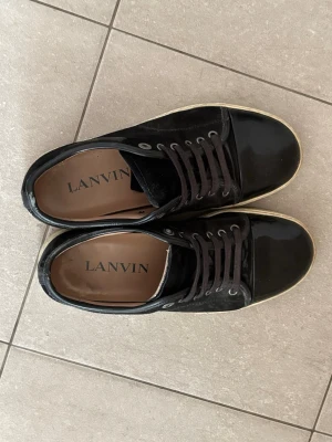 Svarta Lanvin sneakers i mocka och lack - Snygga svarta sneakers från Lanvin med ovandel i mocka och tåparti i blankt lack. Skorna har svarta skosnören, silverfärgade öljetter och en vit sula. Perfekta för dig som gillar stilrena och exklusiva sneakers med en lyxig känsla.
