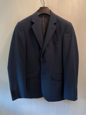 Mörkblå slim fit kavaj 46 - Stilren mörkblå kavaj från Dressmann i slim fit-modell. Kavajen har klassisk passform med två fickor fram, bröstficka och knäppning med en knapp. Insidan är fodrad med randigt tyg och röda detaljer. Perfekt för en dressad look.