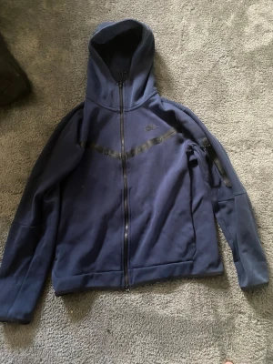 Mörkblå Nike hoodie med dragkedja - Snygg mörkblå hoodie från Nike med hel dragkedja och huva. Har svarta detaljer i form av en diagonal linje över bröstet och Nike-logga på vänster sida. Tillverkad i mjukt material, perfekt för en sportig och avslappnad stil.