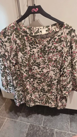 Blommig blus från H&M, storlek 40 - Snygg blus från H&M med blommigt mönster i grönt och rosa på ljus botten. Blusen har rund halsringning och långa ärmar. Perfekt för dig som gillar naturinspirerade prints och en avslappnad men trendig look.