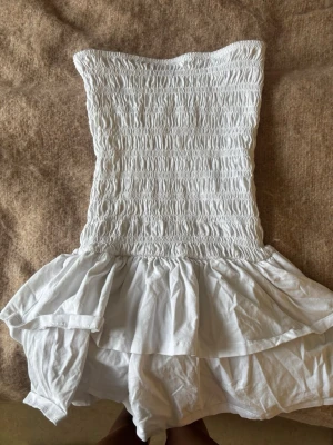 Vit axelbandslös klänning Gina Tricot - Säljer en söt vit axelbandslös klänning från Gina Tricot i storlek XS. Klänningen har smockad överdel och volangig kjol, vilket ger en feminin och somrig look. Perfekt för varma dagar och lätta outfits.