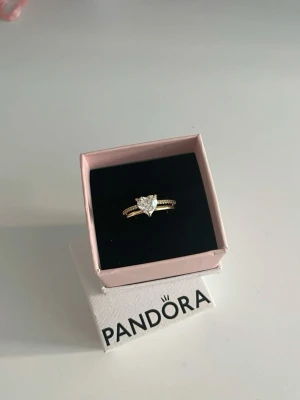 Pandora guld ring med hjärtformad sten - Snygg guld ring från Pandora med en stor hjärtformad klar sten i mitten och små gnistrande stenar längs bandet. Perfekt för dig som vill ha något extra i din smyckessamling.