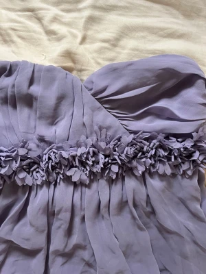 Ljuslila axelbandslös kortklänning - Söt axelbandslös kortklänning i ljuslila med draperad överdel och dekorativa blommor i midjan. Klänningen har en ballongformad nederdel och är tillverkad i ett lätt, flowy material som ger en romantisk känsla.