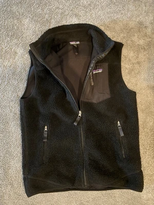 Svart fleeceväst från Patagonia - Svart fleeceväst från Patagonia med dragkedja framtill, två fickor med dragkedja och en bröstficka med logga. Västen har hög krage och är tillverkad i mjukt teddyfleece-material. Perfekt för lager-på-lager och chill stil. Storlek L. Pris kan diskuteras i snabb affär😁