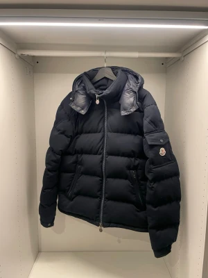 Svart dunjacka från Moncler - Snygg svart dunjacka från Moncler montgenevre med huva och dragkedja framtill. Jackan har två fickor med dragkedja och en ärmficka med Moncler-logga. Puffig modell som håller dig varm under kalla dagar. Perfekt för dig som vill ha både stil och funktion. Kvitto finns tyvärr inte.