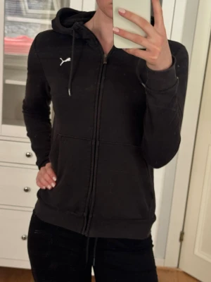 Svart zip hoodie från Puma - Säljer en svart hoodie från Puma med dragkedja framtill och vit Puma-logga på bröstet. Hoodien har huva med dragsko och två fickor framtill. 