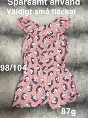 Rosa jumpsuit med enhörningar - Supergullig rosa jumpsuit med volangärmar och elastisk midja. Hela plagget är täckt av ett mönster med vita enhörningar och lila man. Perfekt för dig som älskar färgglada och lekfulla plagg!🌸 Sparsamt använd. Väldigt små fläckar. Storlek 98/104