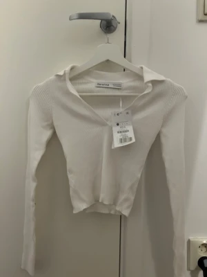 Vit ribbad topp från Bershka - Snygg vit ribbad topp från Bershka med v-ringning och lång ärm. Modellen är croppad och har en stilren design som passar perfekt till jeans eller kjol. Materialet är mjukt och stretchigt för en bekväm passform.