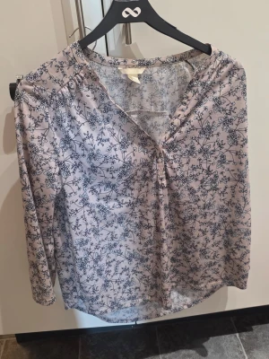 Blommig blus från H&M i ljusblått - Söt blus från H&M i ljusblått med mörkblått blommönster. Blusen har v-ringning och långa ärmar, perfekt för en avslappnad men ändå stilren look. Tunt och lätt material som känns skönt mot huden.