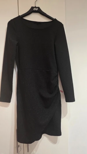 Svart glittrig klänning med lång ärm - Snygg svart glittrig klänning med lång ärm och lätt rynkade detaljer på ena sidan. Klänningen har en kroppsnära passform och är i kort modell, perfekt för dig som gillar stilrena och trendiga plagg.