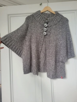 Grå stickad poncho från Mangun - Säljer en snygg gråmelerad stickad poncho från Mangun med bred polokrage och fyra stora dekorativa knappar framtill. Ponchon har en lös och bekväm passform, perfekt att slänga på sig över en tröja. Materialet är mjukt och värmande. Passar allt från xs-m