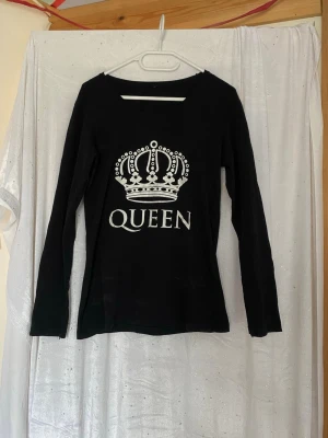 Svart långärmad topp med QUEEN-tryck - Svart långärmad topp med vitt tryck av en krona och texten QUEEN på bröstet. Rak passform och rund halsringning. Perfekt för dig som vill sticka ut med en cool statement-top. Har lite fläckar men det syns bara på nära håll 
