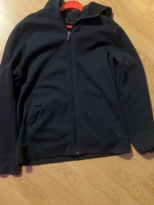 Svart hoodie från Hugo BOSS med dragkedja - (Pris kan diskuteras) Svart hoodie från BOSS med dragkedja framtill och två fickor. Klassisk design med huva och diskret BOSS-logga på bröstet. Perfekt för en chill och stilren look. Materialet känns mjukt och bekvämt mot huden.