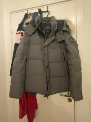 Grå dunjacka från Canada Goose - Snygg grå dunjacka från Canada Goose med avtagbar huva och pälskant. Jackan har en klassisk quiltad design, hög krage och logotyp på ärmen. Perfekt för kalla vinterdagar tack vare det varma dunfodret och den stilrena looken.