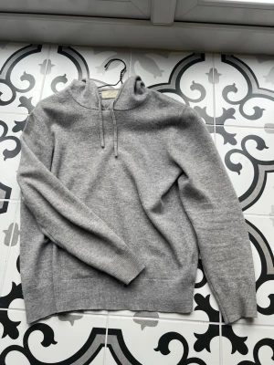 Ljusgrå hoodie i mjuk ullmix - Snygg ljusgrå hoodie med dragsko i huvan och ribbade muddar. Tillverkad i en mjuk och skön ullmix som passar perfekt för en avslappnad stil. Klassisk passform och enkel design gör den lätt att matcha med andra plagg. Defekt vid armhålan