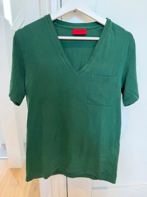 Hugo Boss mörkgrön t-shirt V-ringad stl S – kanonskick - Säljer en stilren och tidlös mörkgrön t-shirt från Hugo Boss i storlek S. Modellen har snygg V-ringning samt praktisk bröstficka som ger en exklusiv och avslappnad look.  T-shirten är i kanonskick och väl omhändertagen. Perfekt både till jeans, chinos eller shorts för en stilren vardagsoutfit.  ✔ Storlek S ✔ Färg: mörkgrön ✔ V-ringning & bröstficka ✔ Kanonskick ✔ Djur- och rökfritt hem  Skickas gärna eller hämtas enligt överenskommelse. Först till kvarn!