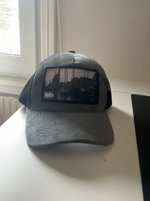 Grå och svart keps med tryck - Snygg keps i grått och svart med mesh på sidorna och baksidan. Framtill finns ett svartvitt tryck med stadsmotiv och texten 'Soulless'. Perfekt för dig som gillar streetstyle och vill ha en unik accessoar.