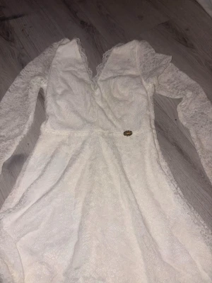 Vit lång spetsklänning med v-ringning - Romantisk vit långklänning med v-ringning och långa ärmar i spets. Klänningen har ett feminint snitt och fina spetsdetaljer över hela plagget. Perfekt för speciella tillfällen när du vill känna dig extra elegant.