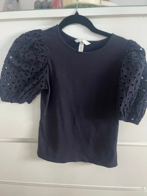 Svart topp med puffärmar från H&M - Supersnygg svart topp från H&M med puffiga, broderade kortärmar i bomull. Ärmarna har hålmönster som ger en cool och trendig look. Perfekt att styla med jeans eller kjol för en extra touch.