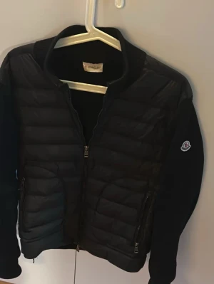 Svart quiltad moncler jacka  - Snygg svart pufferjacka från Moncler med quiltad front och ärmar i mjukt tyg. Jackan har dragkedja framtill, två fickor med dragkedja och klassisk Moncler-logga på ärmen. Perfekt för kyliga dagar och ger en stilren look.
