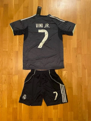 Vinicius Jr Real Madrid matchställ borta - Snyggt svart matchställ från Real Madrid med Vini Jr. och nummer 7 på ryggen. Tröjan har vita detaljer, klubbmärke och sponsortryck framtill. Shortsen har klubbmärke och nummer 7 samt klassiska Adidas-ränder på sidan. Perfekt för fotbollsträning eller match.