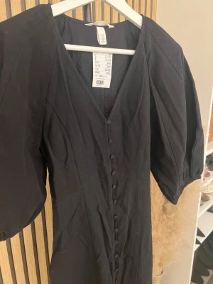 Svart långklänning med puffärm H&M - Svart långklänning från H&M med v-ringning, puffiga kortärmar och knappar längs hela framsidan. Klänningen har en elegant och tidlös design som passar perfekt för dig som gillar stilrena plagg med en twist. Skriv om ni vill ha bild på 🫶🏼🫶🏼