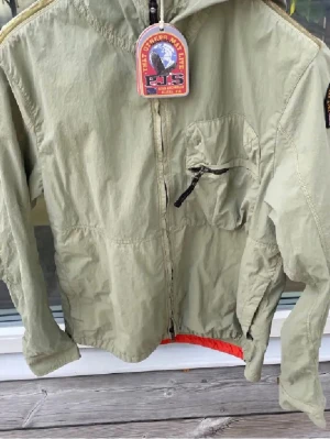 Ljusgrön vindjacka från Parajumpers - Stilren ljusgrön vindjacka från Parajumpers med huva, dragkedja framtill och bröstficka med svart dragkedja. Jackan har en orange detalj på ärmen och patch med logga. Lätt material, perfekt för blåsiga dagar.
