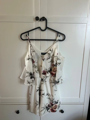 Vit blommig byxdress från Parisian - Supersöt vit byxdress med blommigt mönster i vinrött, rosa och grönt. Smala axelband, v-ringning och volangdetaljer vid axlarna. Lätt och luftig modell, perfekt för varma dagar. Materialet känns mjukt och flowy.