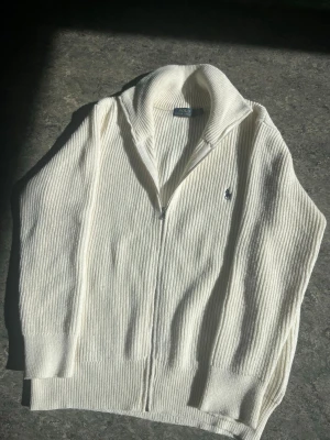Vit ziptröja från Polo Ralph Lauren - Klassisk vit ribbstickad ziptröja från Polo Ralph Lauren med hög krage och dragkedja framtill. Tröjan har det ikoniska broderade logotypen på bröstet och är tillverkad i bomull. Perfekt för en stilren och avslappnad look.