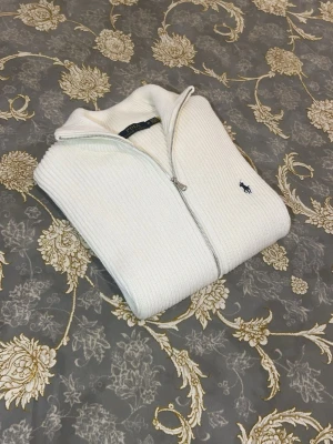 Vit stickad ziptröja från Polo Ralph Lauren - Säljer en vit stickad tröja från Polo Ralph Lauren med silverfärgad dragkedja och klassisk broderad logga på bröstet. Tröjan har ribbad struktur och hög krage, perfekt för att hålla värmen. Enkel och stilren design som passar till många outfits.