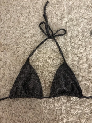 Glittrig svart trekantsbikini - Snygg trekantsbikini i svart med glittrigt material och knyt i nacken. Perfekt för dig som vill sticka ut på stranden eller vid poolen. Enkel och trendig design som passar till sommarens alla bad.