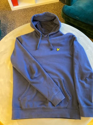 Blå hoodie från Lyle & Scott - Snygg blå hoodie från Lyle & Scott med klassisk gul logga på bröstet. Tillverkad i ekologisk bomull, har huva med dragsko och en stor magficka. Perfekt för en avslappnad och stilren look.