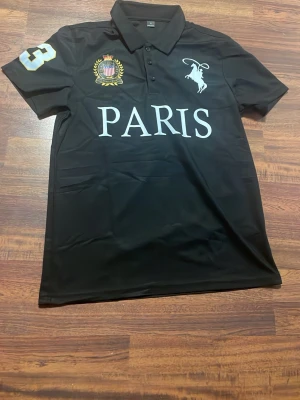 Svart Polo Ralph lauren tröja med Paris-tryck - Svart polo tröja med krage och knappar framtill. Stort vitt tryck med texten PARIS på bröstet, broderad emblem och siffra 3 på ärmen. Materialet känns som polyester och tröjan har korta ärmar. Perfekt för en sportig och stilren look.