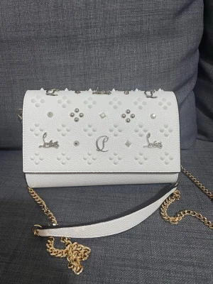 Vit axelväska från Christian Louboutin - Snygg vit axelväska i läder från Christian Louboutin med dekorativa nitar, silverdetaljer och kedjerem i guld. Väskan har präglade mönster och logotyp framtill samt en liten röd detalj på baksidan. Perfekt för att lyfta din outfit.