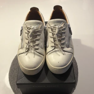 Vita sneakers från Giuseppe Zanotti (lite större i storlek stolek 42 1/2 men passar 43) - Snygga vita sneakers från Giuseppe Zanotti i skinn med rund tå och platt sula. Skorna har snörning framtill och dragkedjor på sidorna för extra edge. Insidan är fodrad i beige och de har diskret logga bak på hälen. Perfekta för dig som gillar stilrena men ändå unika sneakers. Passar storlek 43 (helt äkta)