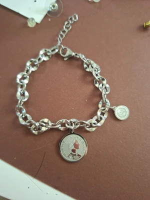 Silverfärgat kedjearmband med myntberlock - Stilrent armband i silverfärgad metall med kraftig kedja och två myntformade berlocker. Ena berlocken har ett motiv av en profil och text runt om. Justerbar längd med karbinlås. Perfekt accessoar för en trendig look.